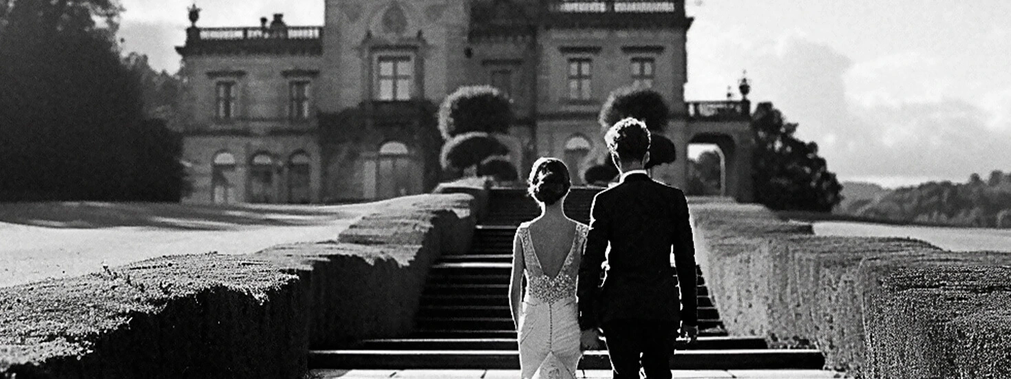 A black-and-white photo of a wedding couple holding hands and walking towards a grand manor, ideal for a classic wedding invitation design.