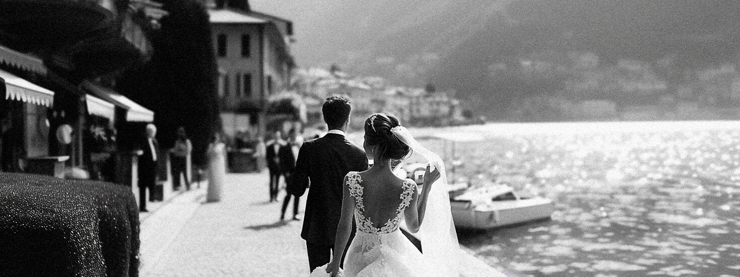 Bride and groom walking by a lakeside in a romantic black-and-white photograph.