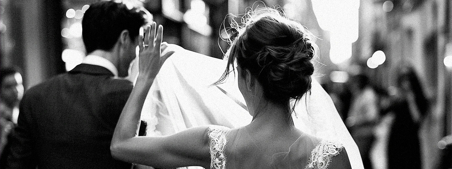 Black and white image of a bride and groom walking away together, with the bride lifting her veil and the groom in a suit.