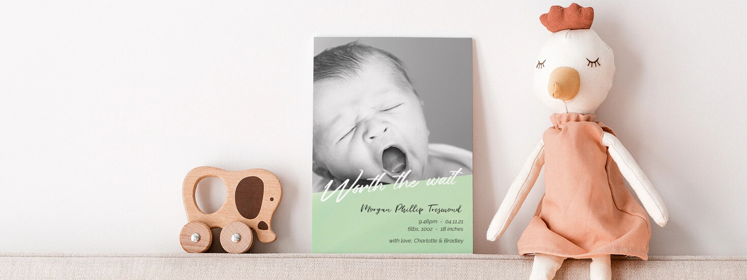 Birth announcement cards displayed on a shelf with soft toys, featuring elegant typography and photo options.