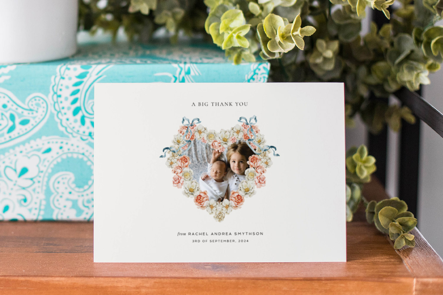 Baby thank you cards with a heart-shaped floral frame featuring two children, displayed on a wooden table with greenery
