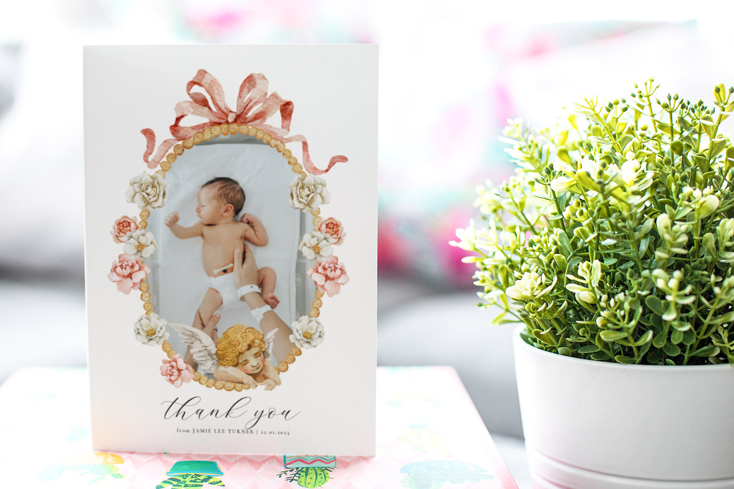 Baby thank you cards featuring a newborn photo framed by floral and angel illustrations, displayed upright beside a green