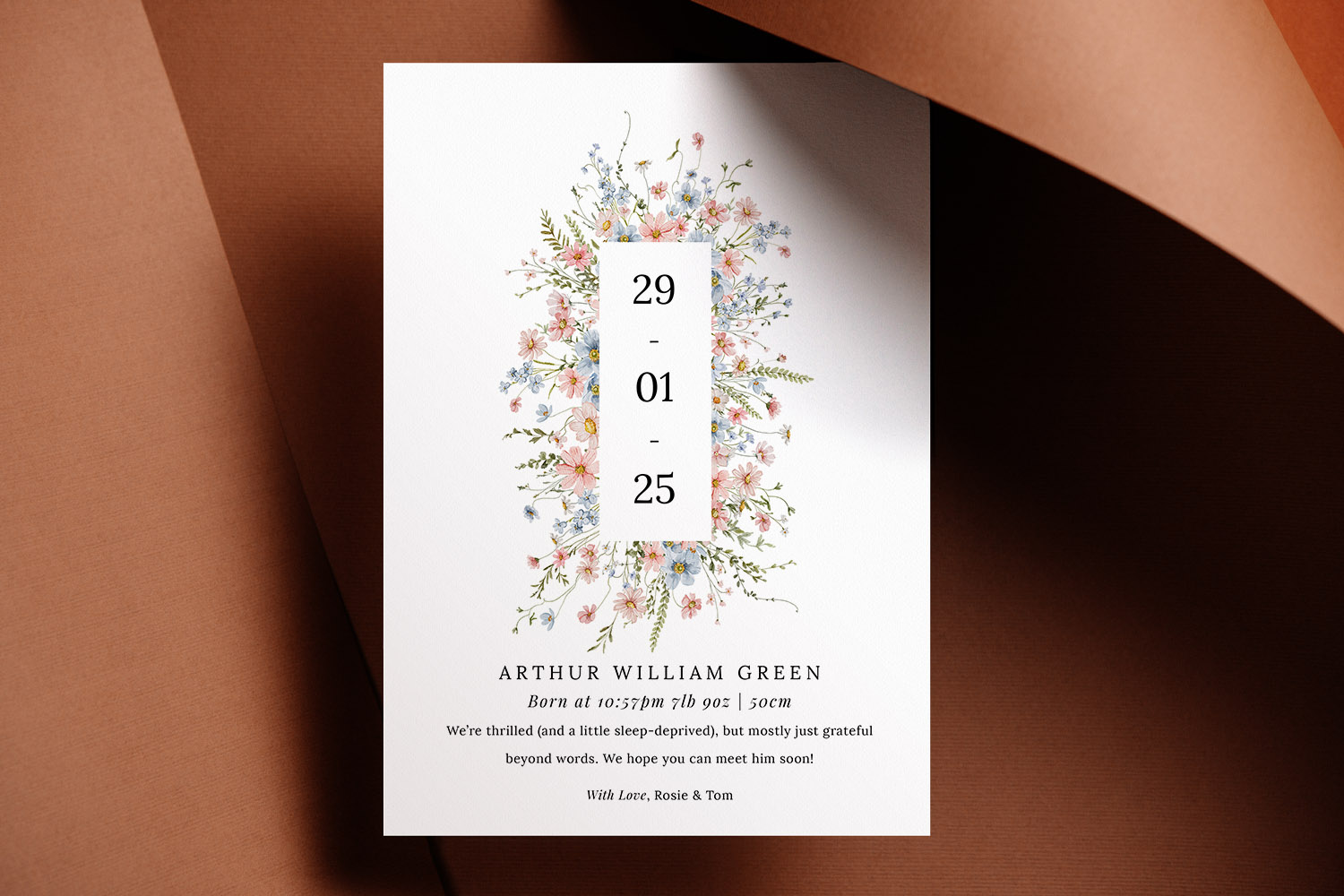 Birth announcement card with delicate floral border and modern typography, displayed on a brown textured background.