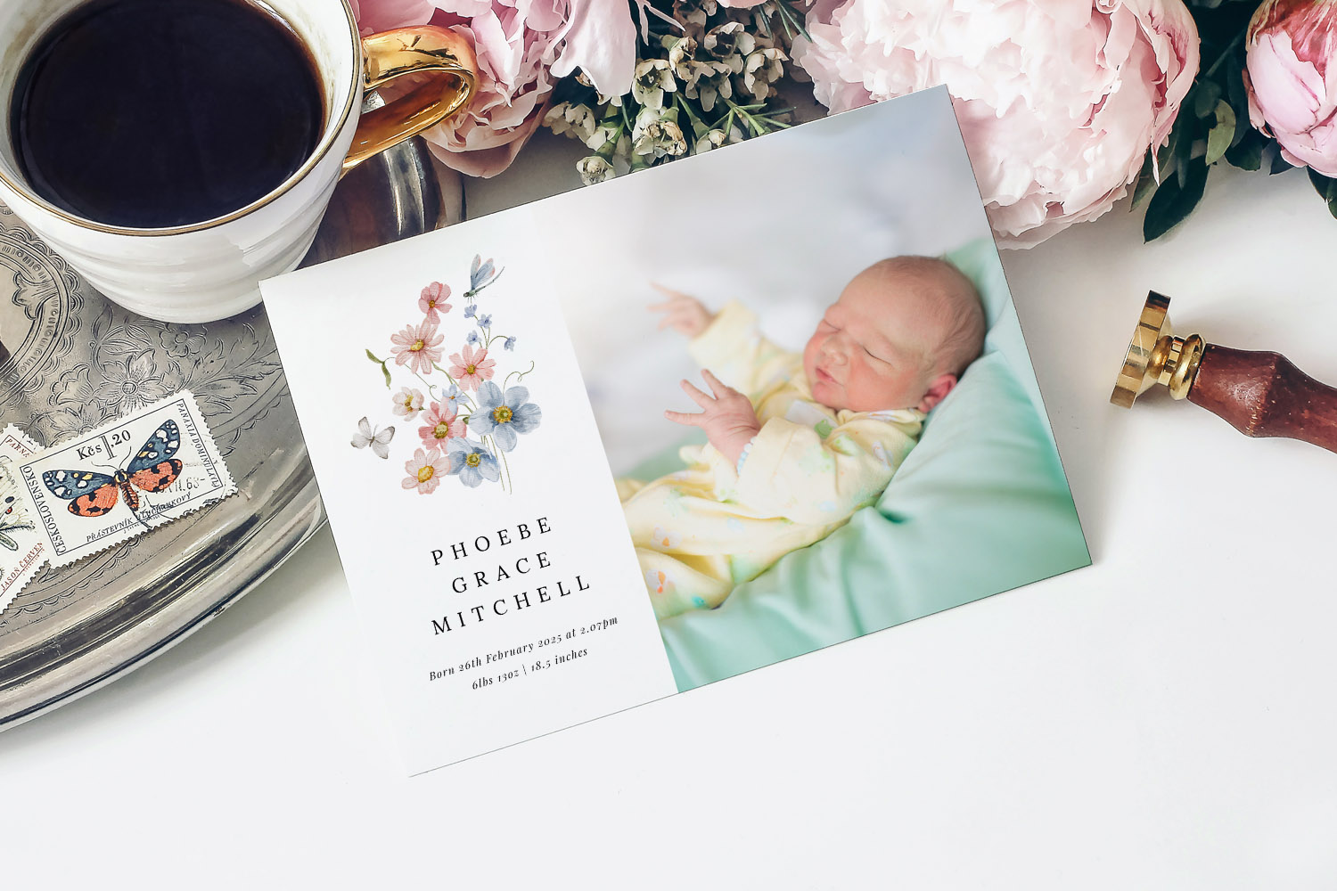 Birth announcement card featuring a floral design and newborn baby photo, displayed on a table with flowers and a coffee cup.