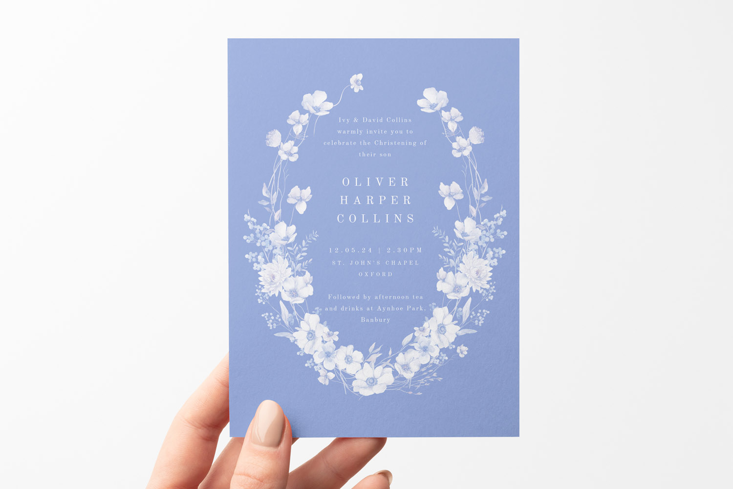 Christening invitations in pastel blue with delicate white floral wreath and elegant serif text, held by a hand against a