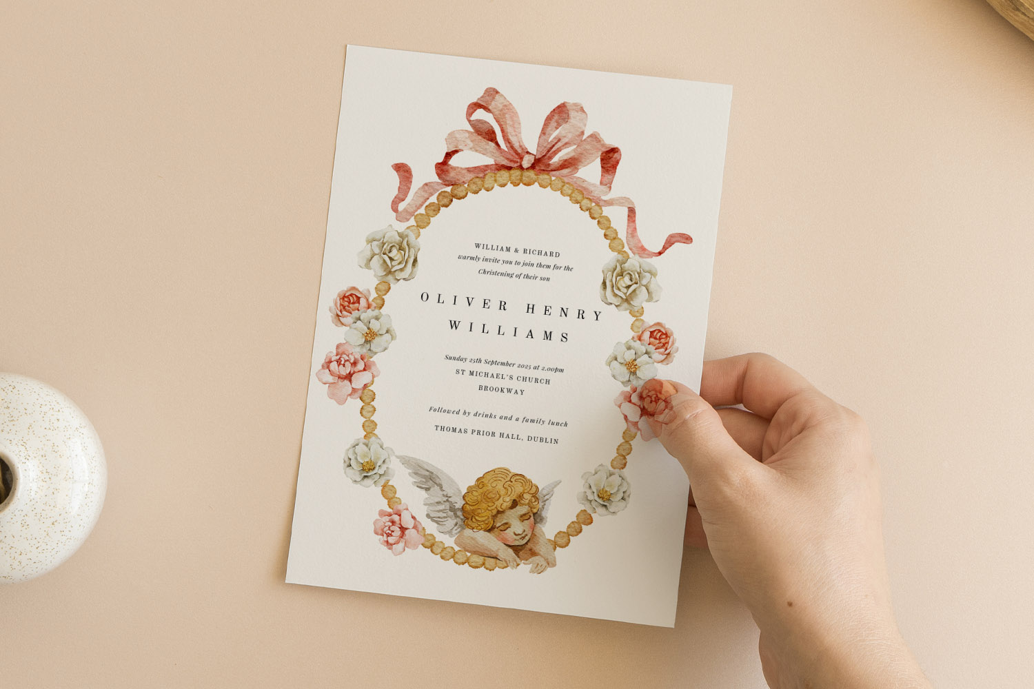 Christening invitations featuring a floral and cherub wreath design with elegant script, held by hand on a neutral