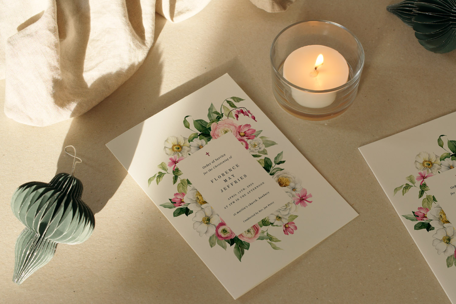 Christening Order of Service card with elegant floral border, displayed on a table beside a lit candle and green ornament.