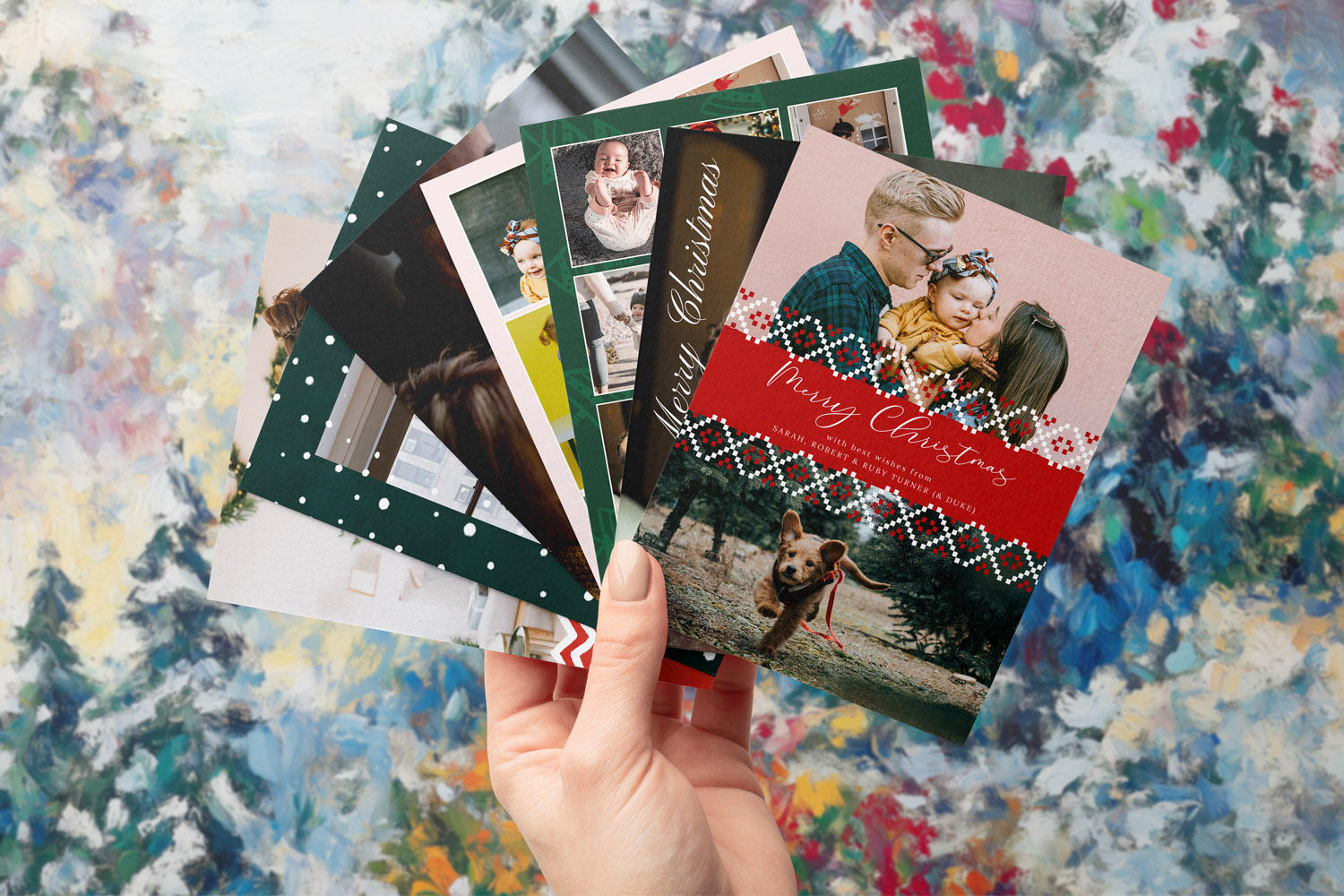 Christmas cards with family photos and festive designs held in a hand against a colourful painted winter background.