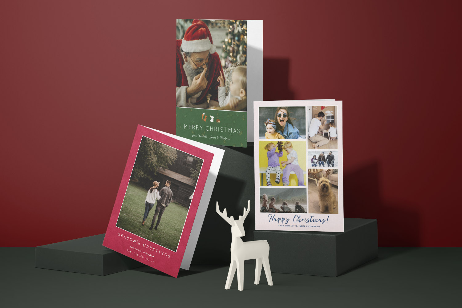 Christmas cards with personalised photo designs displayed on dark stands against a red background with a white reindeer