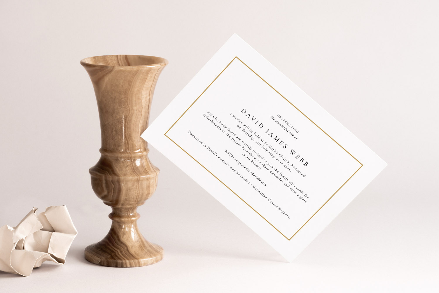 Funeral announcement card with gold border and black text, displayed upright beside a marble vase on a neutral background.