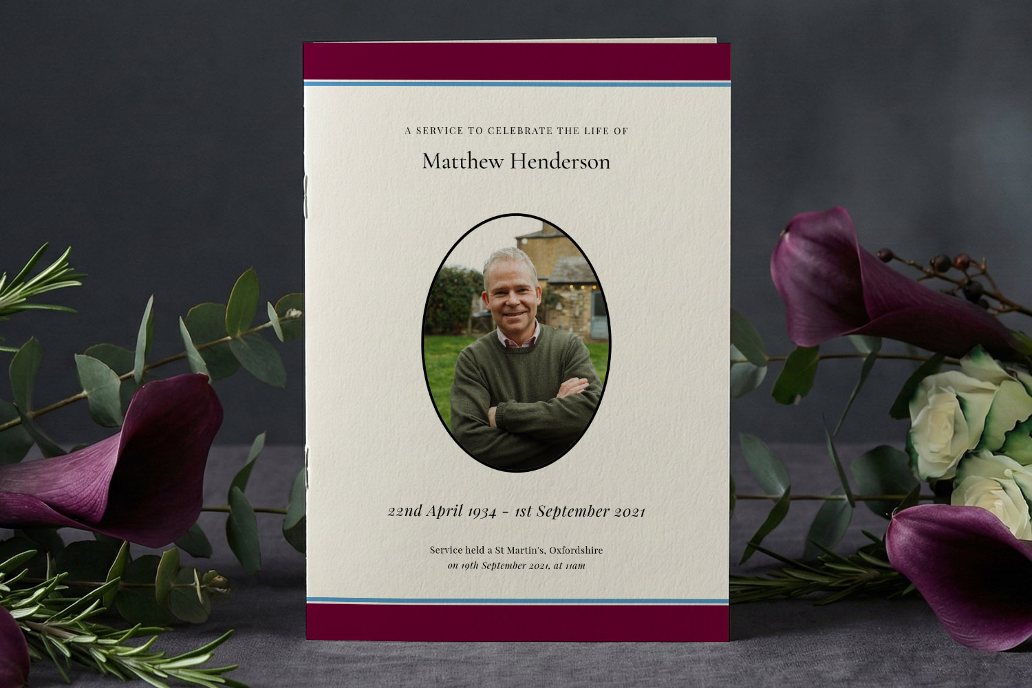 Funeral order of service booklet with a cream cover, maroon border, and an oval photo, displayed with flowers on a dark