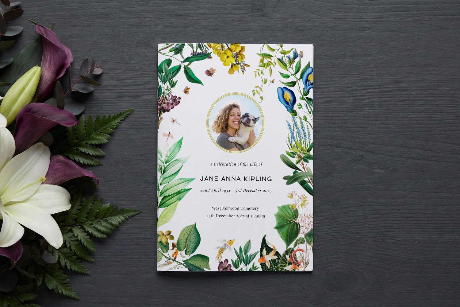 Funeral order of service booklet with floral border and photo, displayed on a dark wood surface beside lilies and greenery.