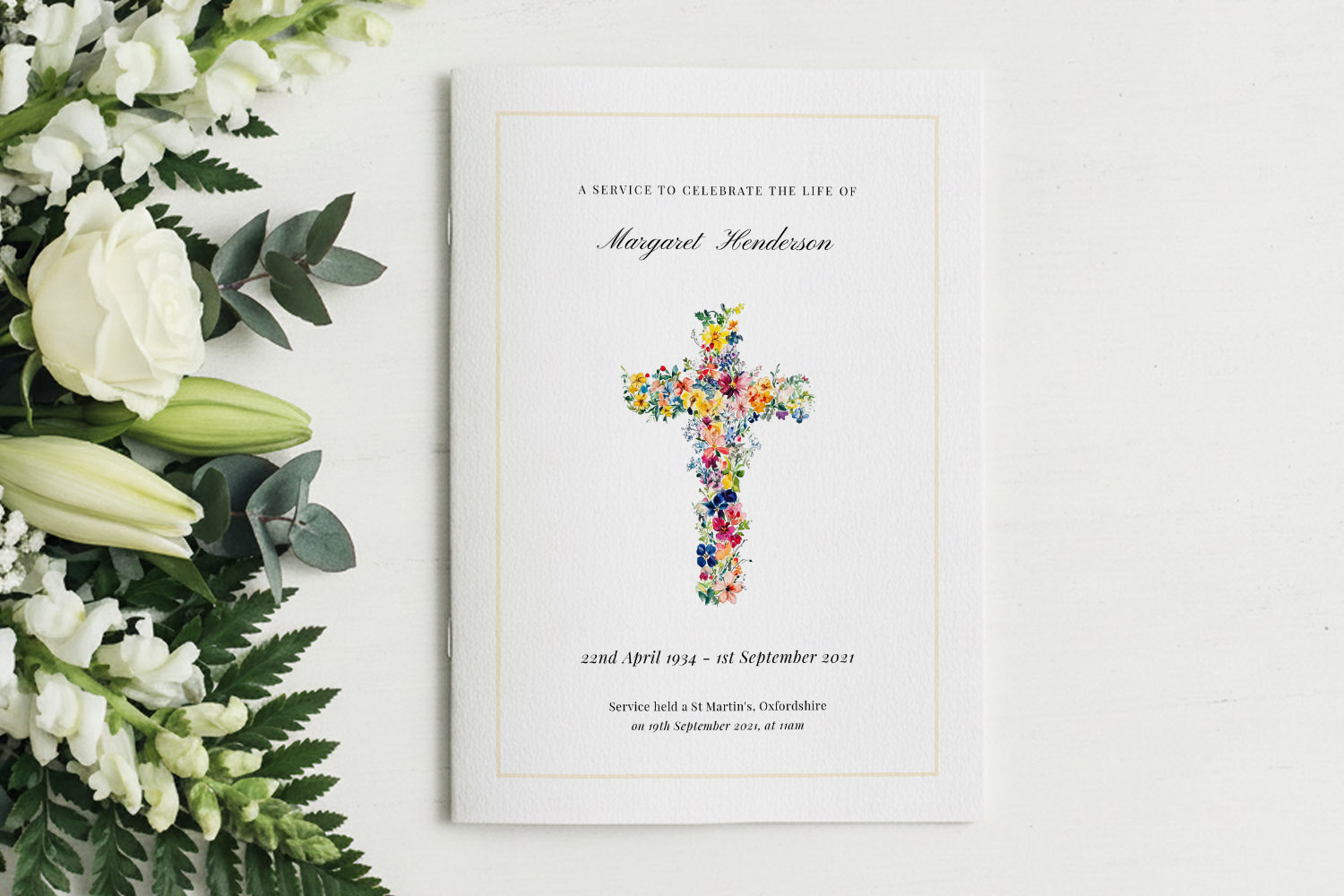 Funeral order of service booklet with a colourful floral cross design on the cover, placed beside white lilies and roses.