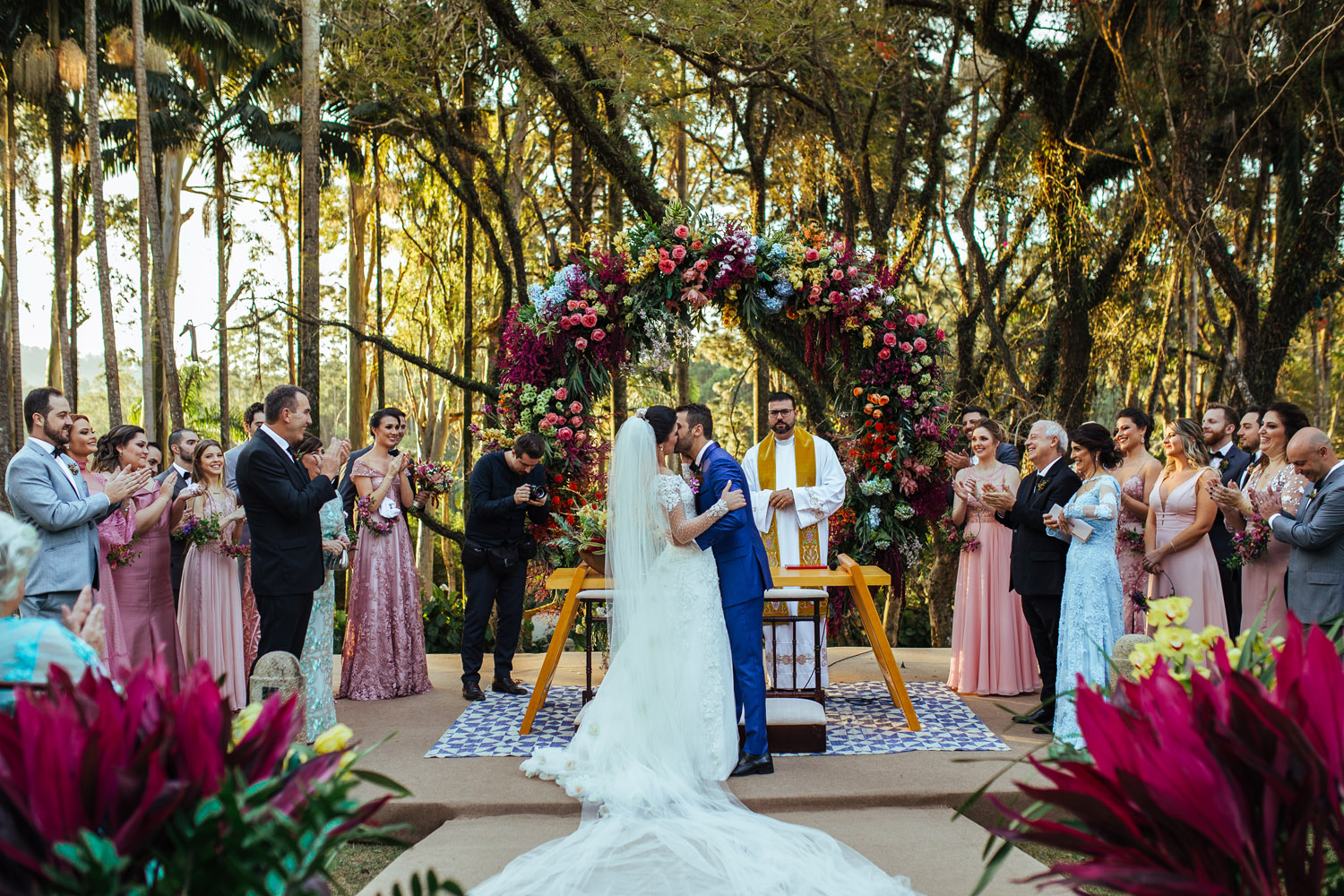 RSVP.studio outdoor wedding ceremony with bride and groom kissing under a vibrant floral arch, surrounded by guests and trees