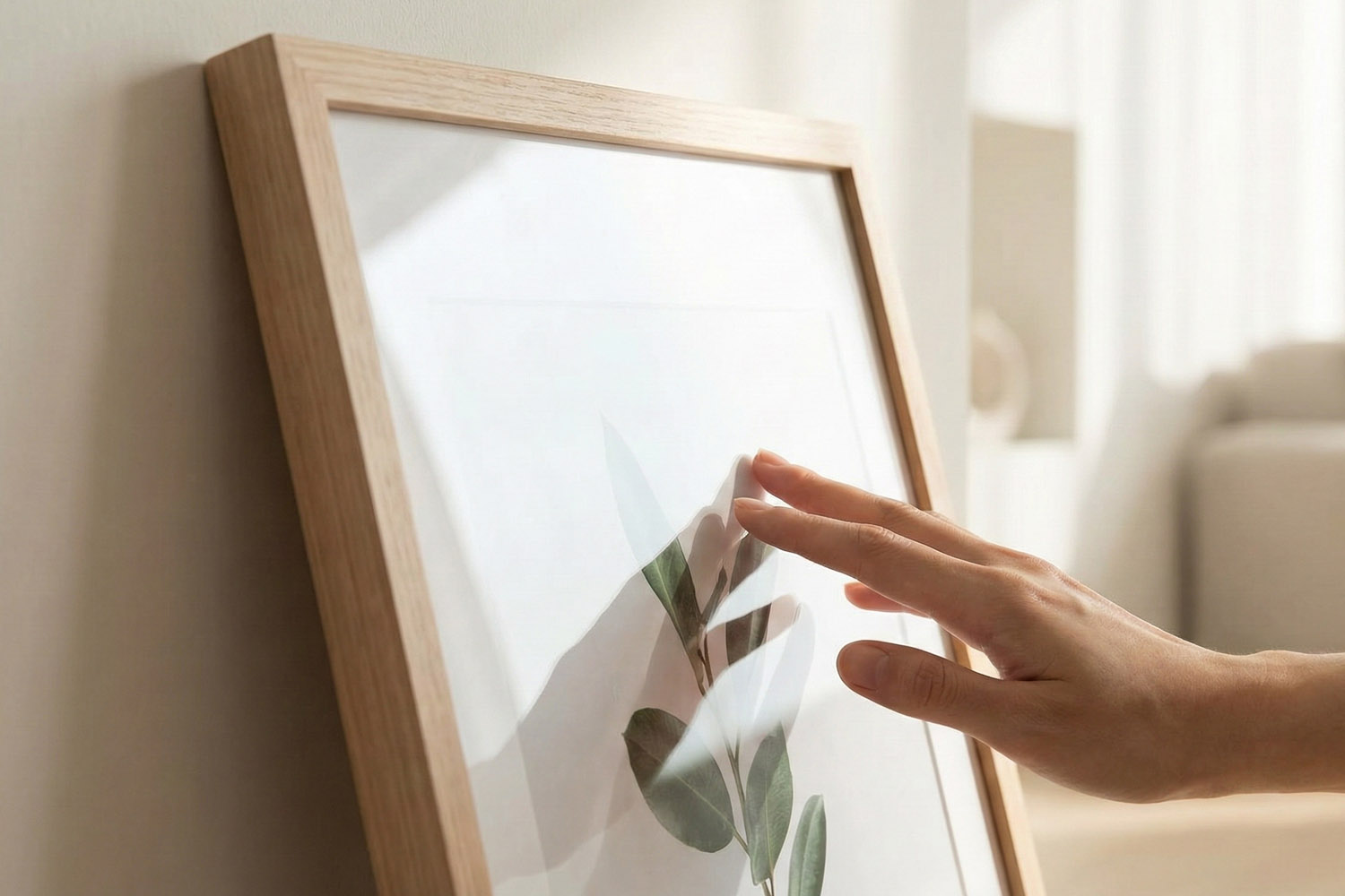 Simple framed prints with a light wood frame and botanical artwork, shown being touched in a softly lit modern interior.