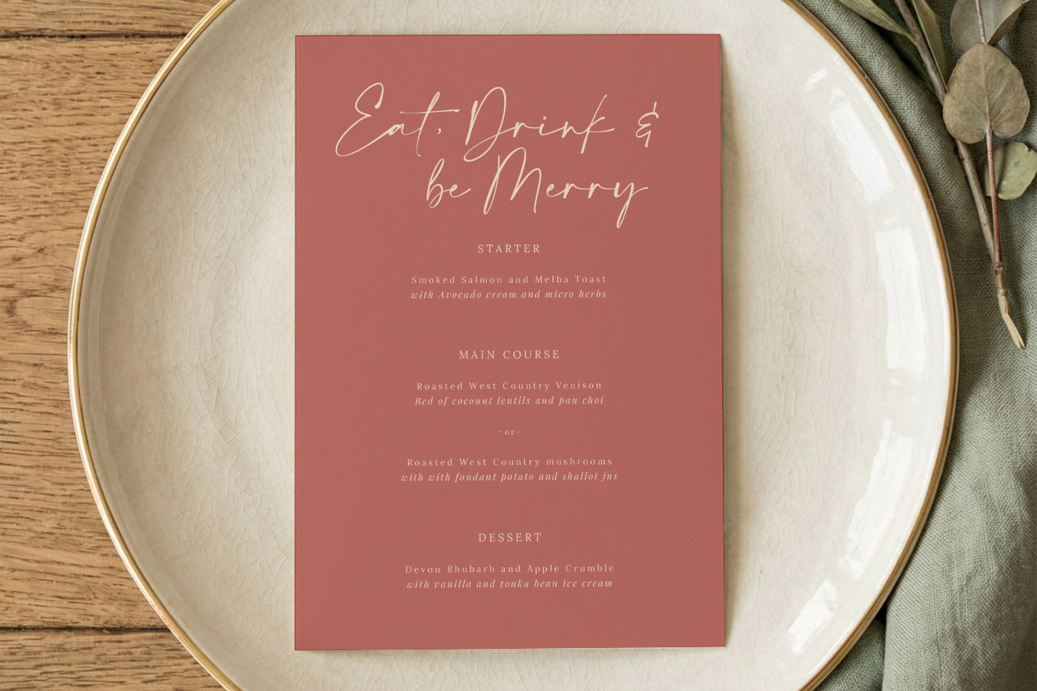 Wedding menu card in blush pink with elegant white script, displayed on a ceramic plate with green napkin and foliage.