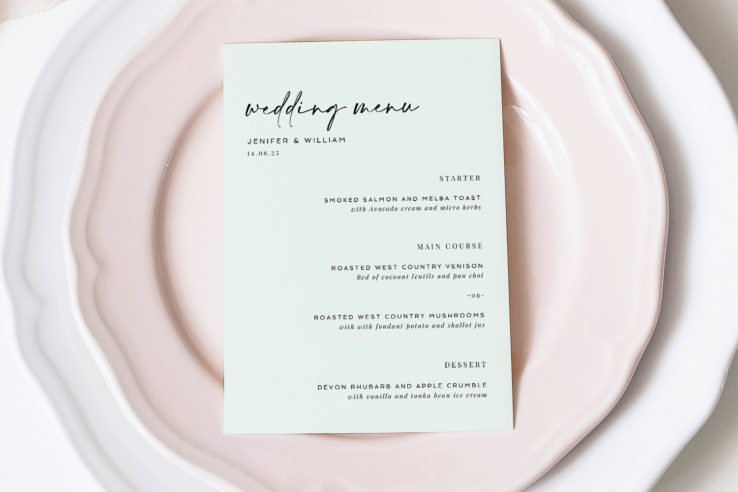 Wedding menus featuring minimalist black script on white card, displayed on layered blush and white plates from a top-down