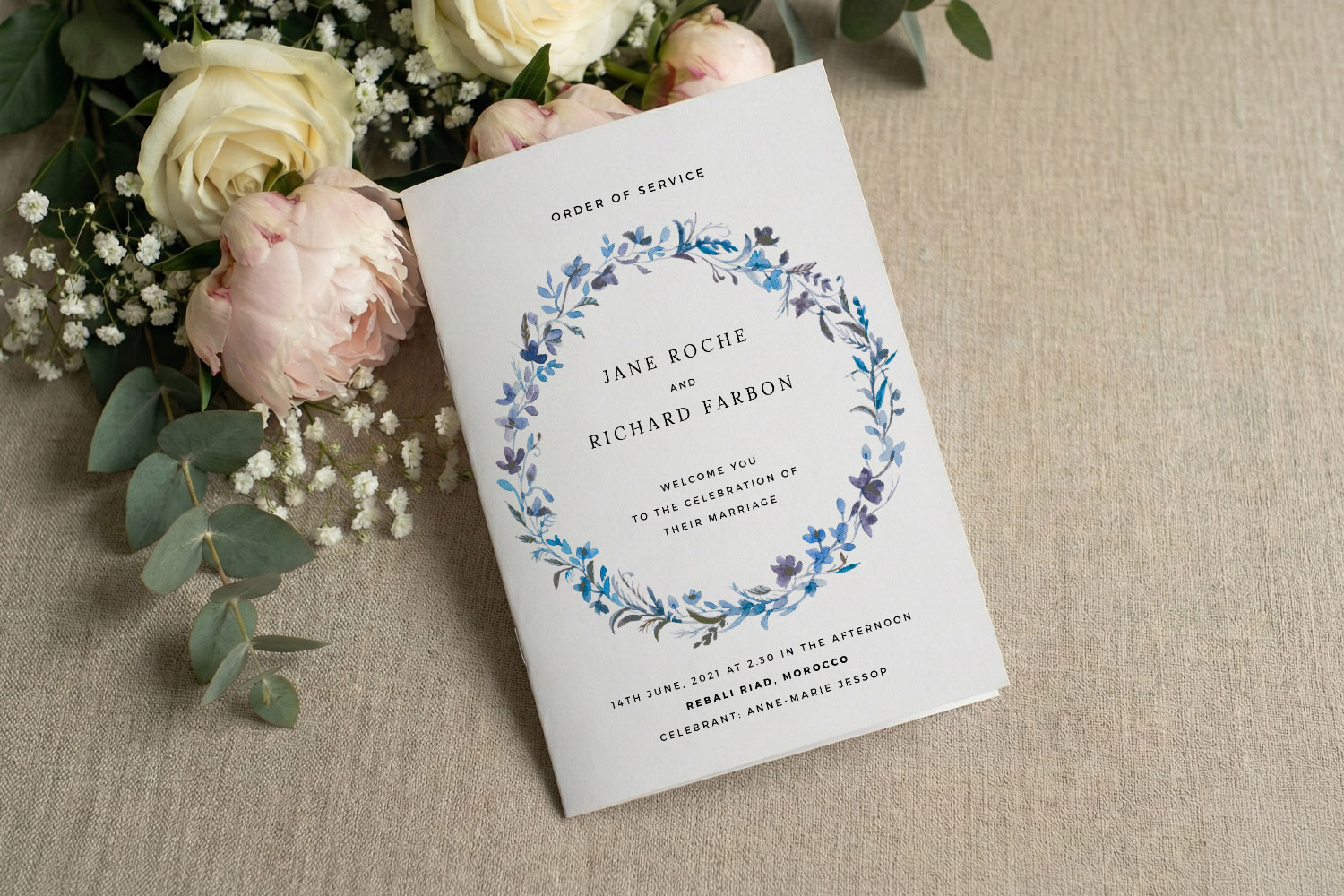 Wedding order of service booklet with blue floral wreath design, displayed on linen with blush roses and greenery beside it