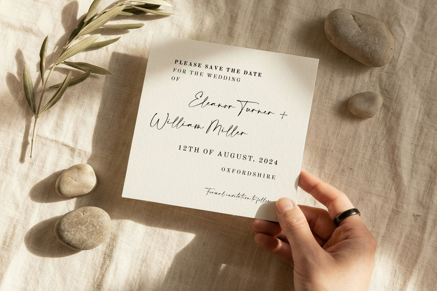 Wedding save the dates card with elegant script font, held by a hand on a linen cloth with pebbles and olive branch.