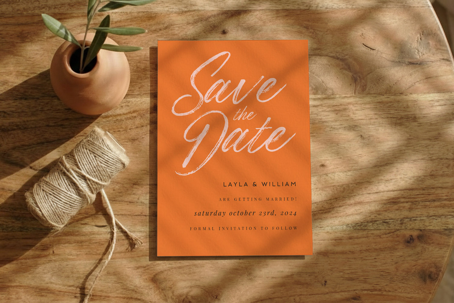 Wedding save the dates card in bold orange with white script lettering, displayed on a rustic wooden table with twine and a