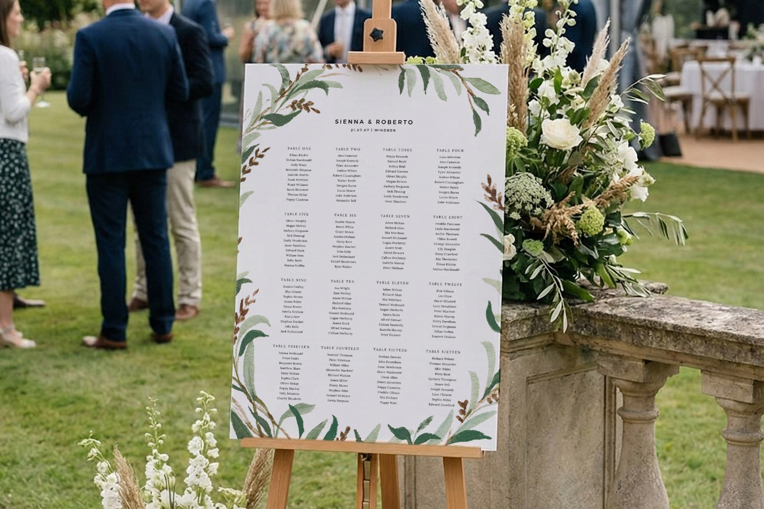 Wedding table plan with botanical border displayed on an easel outdoors, surrounded by floral arrangements and guests.