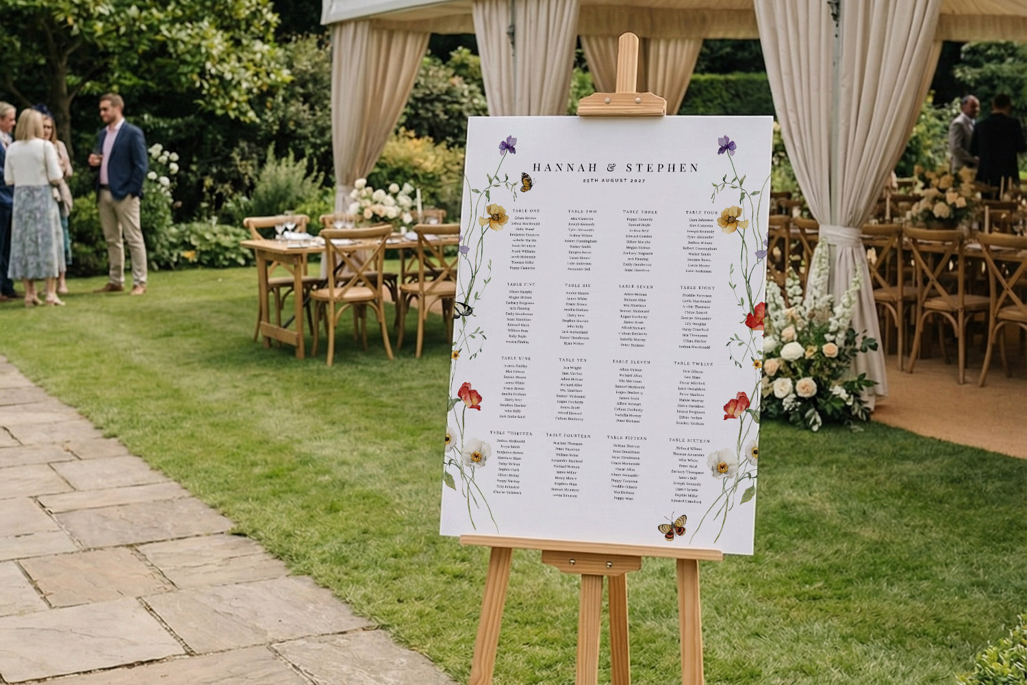 Wedding table plan with floral border on easel, displayed outdoors beside wooden chairs and tables under a marquee.