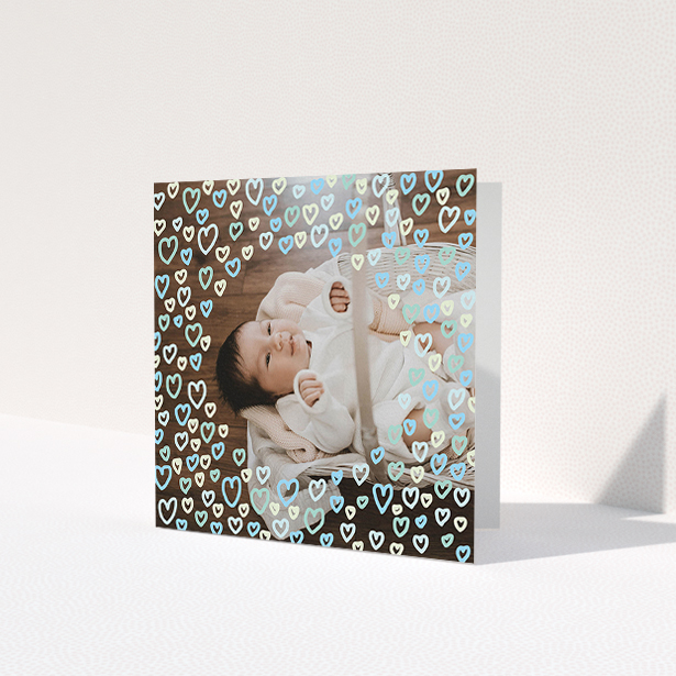 A baby thank you card named "Heart Overlay". It is a square (148mm x 148mm) card in a square orientation. It is a photographic baby thank you card with room for 1 photo. "Heart Overlay" is available as a folded card, with mainly blue colouring.